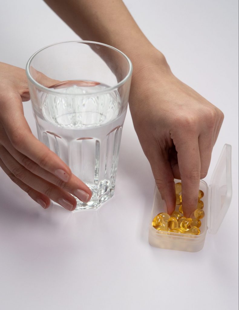 Images shows 2 hands. One is holding a glass of water and the other is reaching for a gelatine capsule from a pill box.